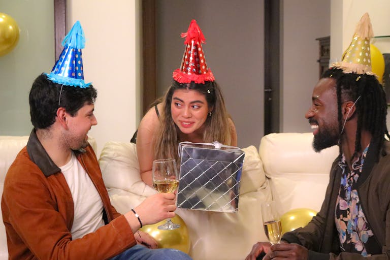 Three friends celebrating a birthday indoors with drinks and party hats.