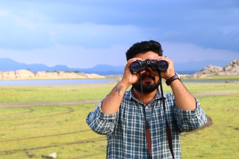 Man with beard and tattoo using binoculars in a grassy field under a blue sky.