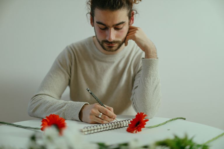 A focused man writing in a notebook, surrounded by red flowers on a table in an indoor setting.