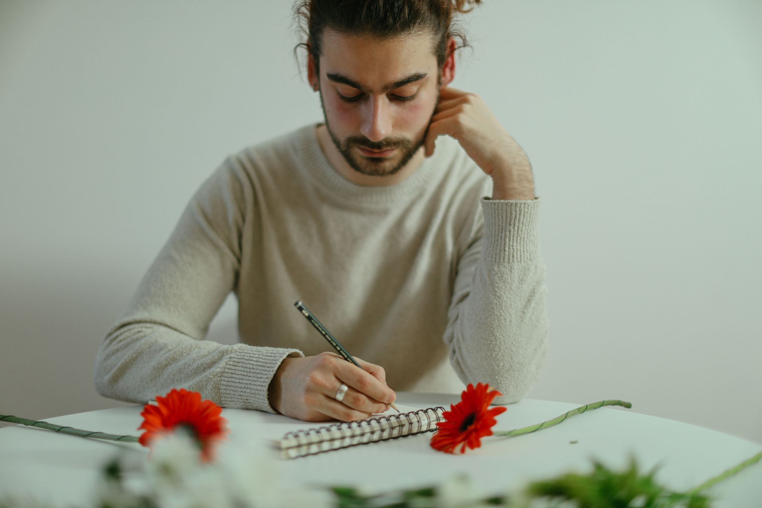 A focused man writing in a notebook, surrounded by red flowers on a table in an indoor setting.