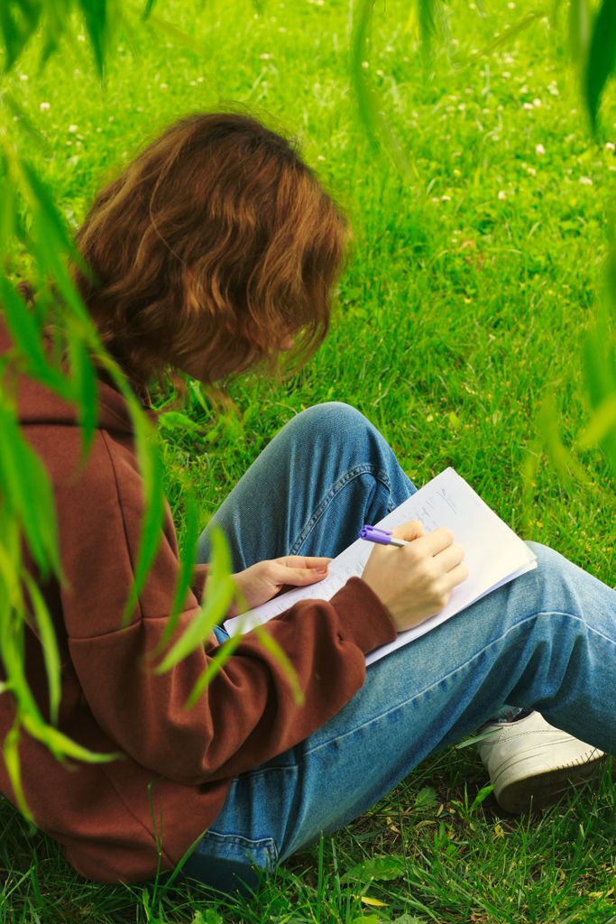 A young adult sits on grass, jotting notes in a notebook, surrounded by lush greenery.