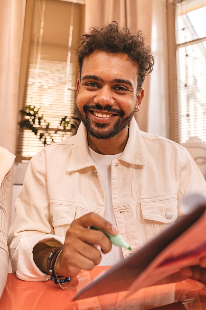 Cheerful man with curly hair highlighting notes while smiling warmly at the camera indoors.
