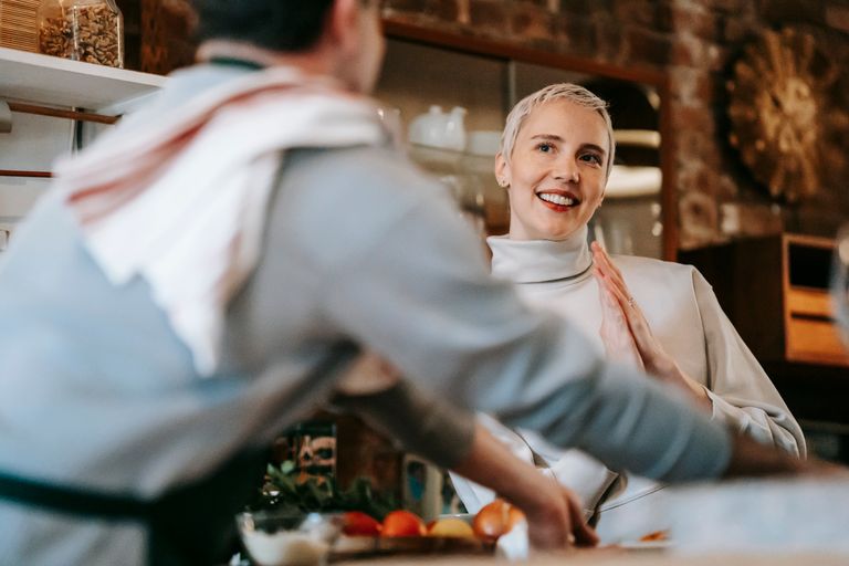 Positive adult couple in bright kitchen while male cooking meal on counter with various ingredients and products near female friend while looking at each other
