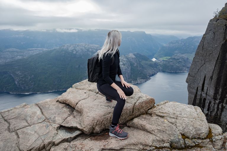 Woman sits on Preikestolen cliff in Norway, overlooking fjord. Explore stunning mountain landscapes.