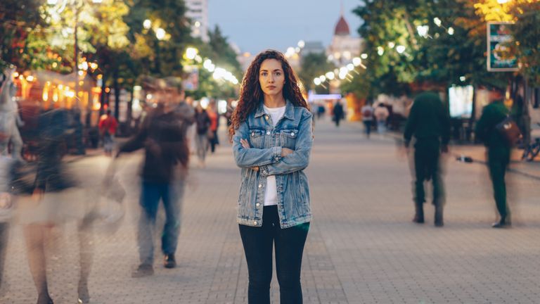Young woman in denim jacket standing calmly on vibrant city street at dusk.