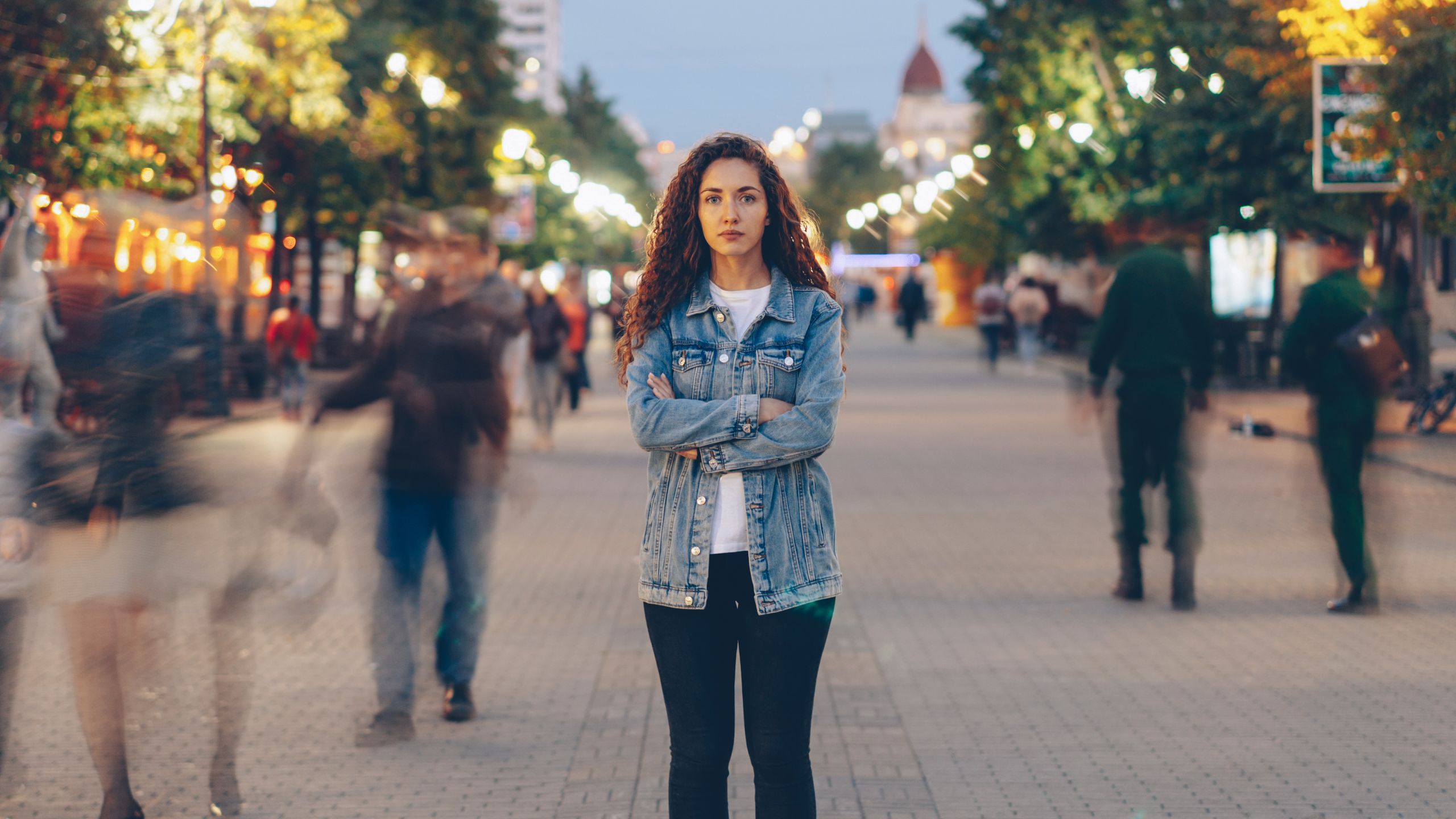 Young woman in denim jacket standing calmly on vibrant city street at dusk.